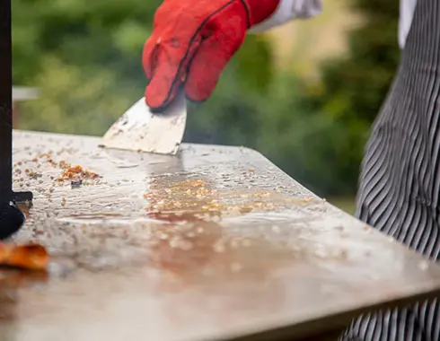 Personne portant des gants et utilisant une sptaule pour nettoyer la plancha d'un brasero
